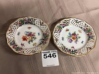 Two reticulated porcelain dishes with colorful floral patterns and gold trimming, placed on cloth.
