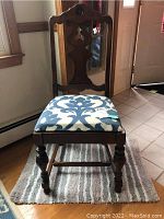 Wooden chair with carved backrest and blue-white patterned upholstered seat on a striped gray-blue-white rug, showing side angle and details of chair legs.