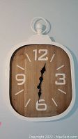 Front view of the battery operated wall clock showing white enameled metal frame, woodgrain face with white numerals and black hands.