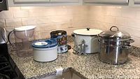 Photo of a kitchen countertop showing all items in the lot: salad spinner, tin with cookie cutters, enamel pot with lid, stainless pot with lid and steamer insert, Brita water pitcher.