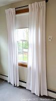 Two white fabric curtain panels hung on a curtain rod over a window with a white roller shade behind.