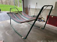 Full view of assembled hammock with green metal frame, striped fabric, and wooden spreader bars, standing on concrete floor.