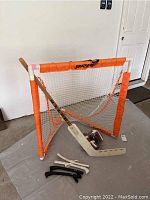 Full view of orange lacrosse net with white netting, wooden lacrosse stick leaning on net, and two skate covers lying in front on floor