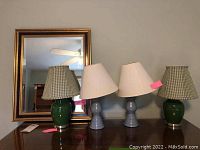 Photo showing four lamps (two green ceramic base with checked shades and two blue wooden base with white shades) arranged in front of the wood framed mirror.
