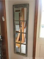 Tall narrow mirror with eight rectangular panes in a distressed hand painted wood frame hung on wall reflecting hallway stairs and door.