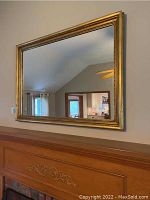 Full view of the rectangular wall mirror hung above a wooden fireplace mantel, showing the gold-toned enamel decorative frame and reflection in the mirror.