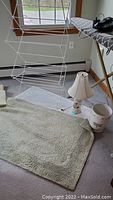 Metal drying rack, ceramic lamp, beige bath rug, ceramic waste basket, and part of an ironing board in room with windows and baseboard heater.