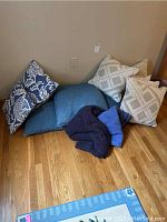 Photo showing a variety of pillows in tan, blue, and white tones arranged on hardwood floor, along with two blankets.