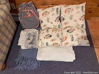 Wide shot showing all items: four aquatic themed pillows, Ame & Lulu backpack, folded blanket, floral cloth, and light bed sheet.