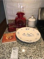 Photo showing ruby red vase, red glass box with bronze hinge, white jar with grape design and lid, blue and white plate and a cookie cookbook on a granite countertop.