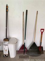 Full overview of lot showing telescoping roof rake in box, two leaf rakes, metal shovel, metal ground rake, and white waste basket