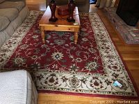 Rug with red center and cream border with floral motifs under a wooden coffee table between couches and near a fireplace