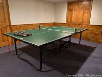 Full view of the green Sportcraft fold-up ping pong table set up in a room with wood paneling and carpet. The paddles and balls are visible on the table.