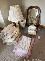 Photo showing ceramic table lamp with pleated shade, wood framed mirror leaning against wall, ceramic waste basket behind, and stacks of folded towels in neutral colors.
