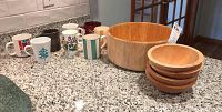 Photo of all items: one large wooden salad bowl, four small wooden salad bowls stacked, and seven ceramic coffee mugs lined up on countertop.