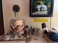 Photo showing a desk surface with assorted office supplies, figurines, and decorative items, including a framed waterfall photo and metal wall hook on the wall above the desk.