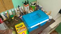 Workbench area with various acrylic paints, craft brushes and a box of Crayola markers.