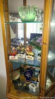Glass cabinet shelf showing collectible car models on boxes, Hummel figurines, a decorative blue-and-white vase with faux flowers, and a green decorative glass bowl with animal glass figurines