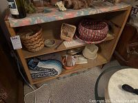 The full bookshelf shown with various baskets and items on the shelves and top. The bookshelf is wooden with a rectangular shape and two shelves. The top is covered with pastel fabric runner.