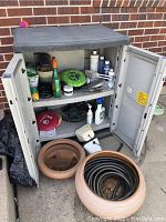 Full view of open plastic storage cabinet with assorted gardening products visible on two shelves; terracotta pots and coiled hose on ground in front.