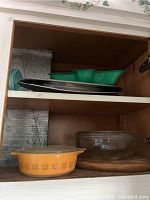 Upper and lower cabinet shelves showing vintage Pyrex orange mixing bowl with lid, green Corelle dish, plates, and other glassware.