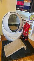 White spherical automatic cat litter box with litter inside, control panel visible, and black-tan carpet mat in front.