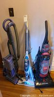 Photo showing three vacuum cleaners and one steam mop arranged side by side on floor with wooden backdrop.