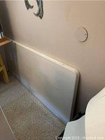 Folded white folding table leaning against wall on carpet floor, shows surface texture and edge shape