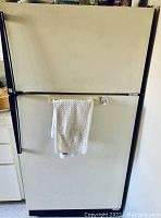 Front view of white Amana refrigerator showing entire unit with top freezer and towel hanging on handle.