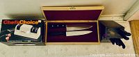 Photo showing the Chef's Choice Diamond Hone Sharpener 110 in original box, two Chicago Cutlery Metropolitan knives in wooden case, and black knife block with knives and scissors.