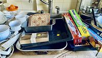 A collection of kitchen bakeware, plastic wrap and wax paper boxes, glass and plastic containers, and cooking pots and pans displayed on a countertop near a sink.