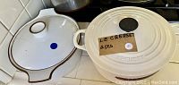 Photo of a white Le Creuset Dutch oven pot with lid and a ceramic pot with brown trim and lid, both used, placed on a stove.