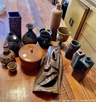 Image showing a collection of pottery vases in various shapes and tones, a lidded container, a ceramic pitcher and cup set, and a carved wooden mask laid out on a wooden surface.