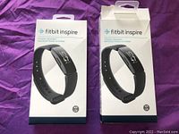 Two Fitbit Inspire fitness tracker boxes side by side on a purple fabric background, one open and one unopened.