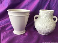 Two white porcelain vases on purple fabric background, showing one smooth tapered vase and one floral embossed vase with handles