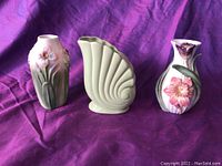 Three petite ornate ceramic vases displayed together, showing different shapes and floral designs on a purple background.