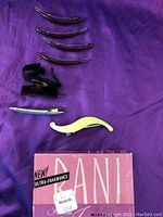 Seven assorted vintage hair clips arranged vertically on purple cloth alongside a Kanebo ultra-fragrance box