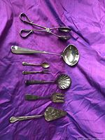 Photo of all seven assorted silver plated serving utensils laid on purple cloth.