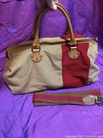 Front view of the oversized bowling bag with two brown leather handles and beige fabric body with a vertical red stripe, alongside the detachable brown and burgundy striped shoulder strap with leather ends.