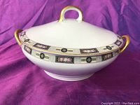 White porcelain round covered serving dish with a decorated band and gold-accented handles on a purple fabric background.