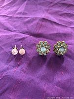 Two pairs of costume stud earrings on a purple fabric background: one small round pink stone pair, one blue and gold floral pair.
