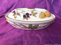 Close-up angle of the covered casserole dish showing fruit and wheat decoration on the lid and body with gold handles.