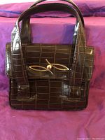 Front view of the dark brown faux crocodile handbag showing the front flap with gold-tone clasp and pearl detail.