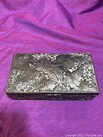 Closed view of hammered metal box with eagle motif on lid and floral accents on sides, placed on purple fabric.