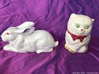 Photo of hand painted ceramic white bunny figurine with red eyes and purple nose detail, next to white ceramic cat figurine with green eyes, red bow tie, and golden paw details on purple fabric background.