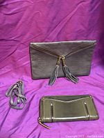 Grey Italian leather clutch with two zipper tassels on front and detached shoulder strap on side, shown on purple background.