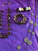 Four jewelry pieces displayed on purple fabric: multi-strand wrap bracelet, stretch bracelet, long necklace with circular pendants, short necklace with large glass beads.