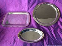 Three large metal serving trays on purple fabric background, one rectangular, one round, one oval.