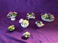 Seven miniature porcelain flower arrangements on purple fabric background showing variety of flower clusters and containers.