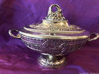 Front view of the ornate silver-colored dish with lid and two handles, displaying detailed stamped biblical imagery and floral motifs.